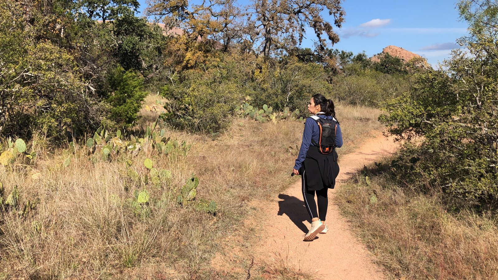 A woman walking a sandy trail through Texas Hill Country brush and prickly pear cactus, with a rocky outcrop rising in the distance