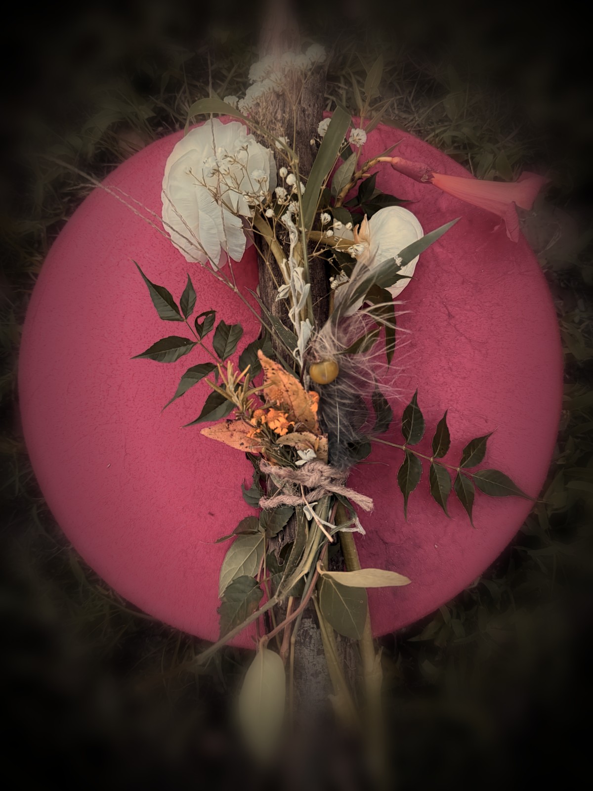 A red shamanic drum resting in the grass, with a hand-tied offering of white flowers, green leaves, feathers and an orange trumpet blossom laid across it