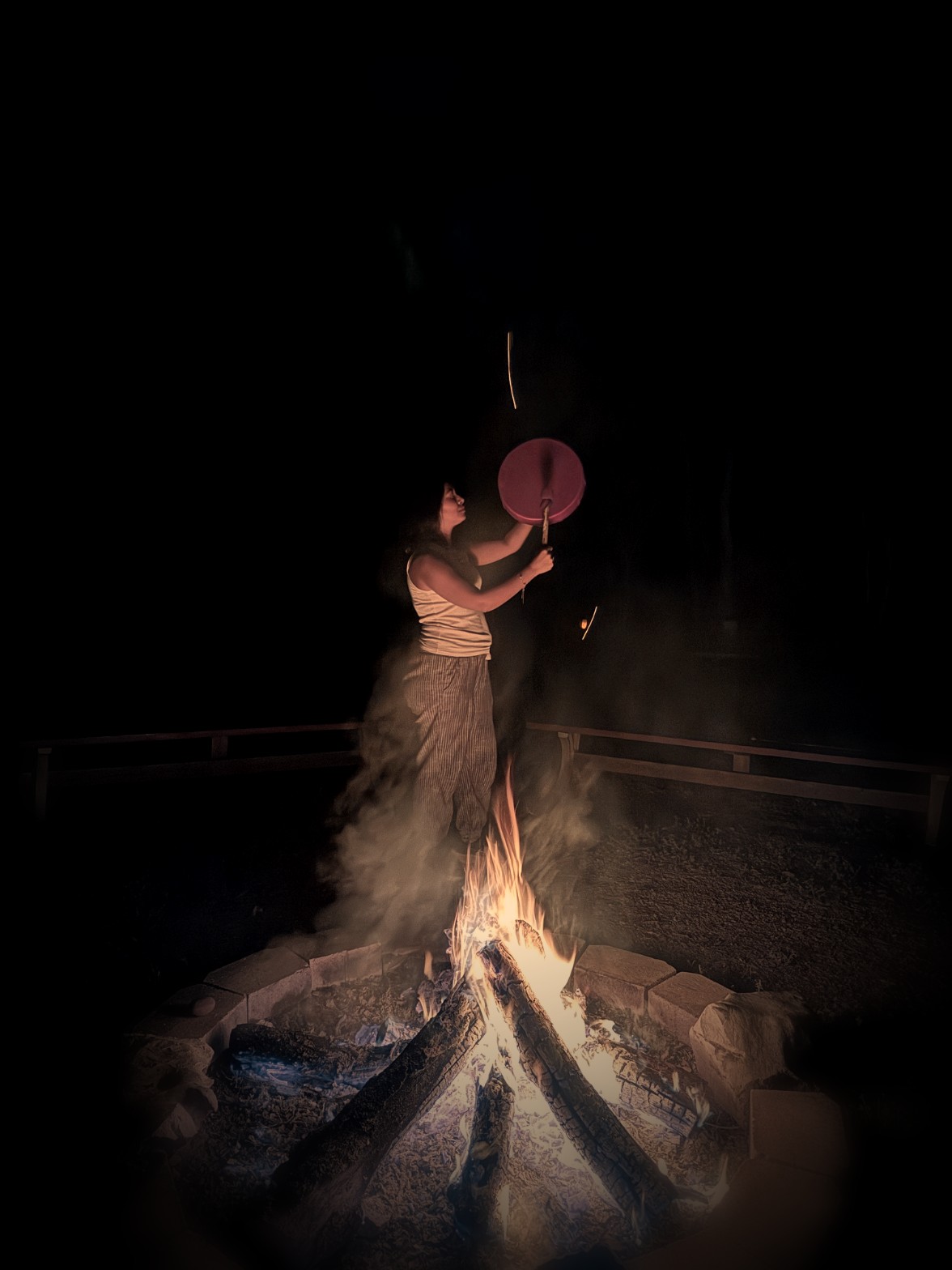 Woman drumming beside a glowing nighttime bonfire in an ancestral healing ceremony